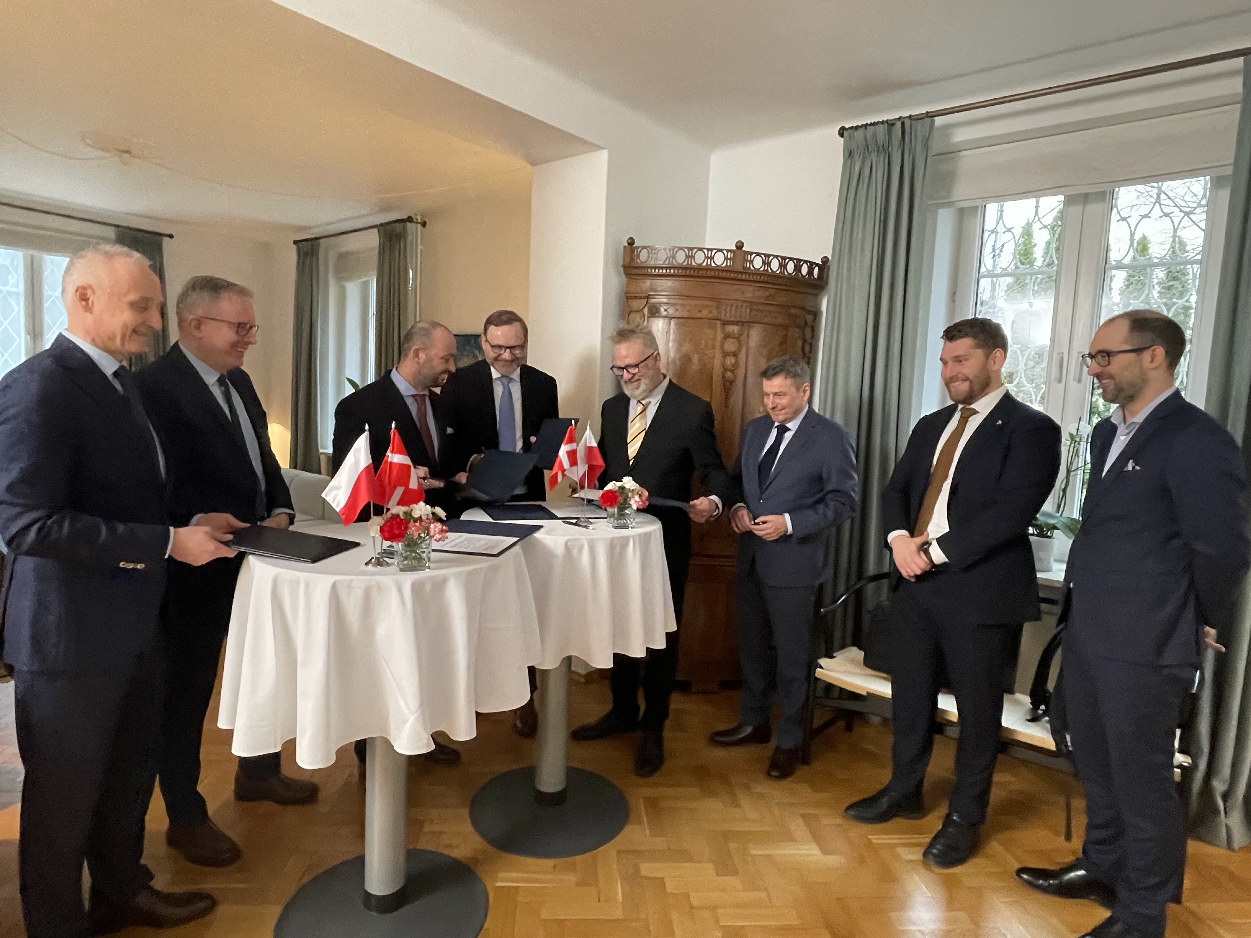 Reprezentanci stron porozumienia stoją przy stołach. Na stołach stoją flagi Polski i Danii oraz  leżą dokumenty. Mężczyźni uśmiechają się do siebie.